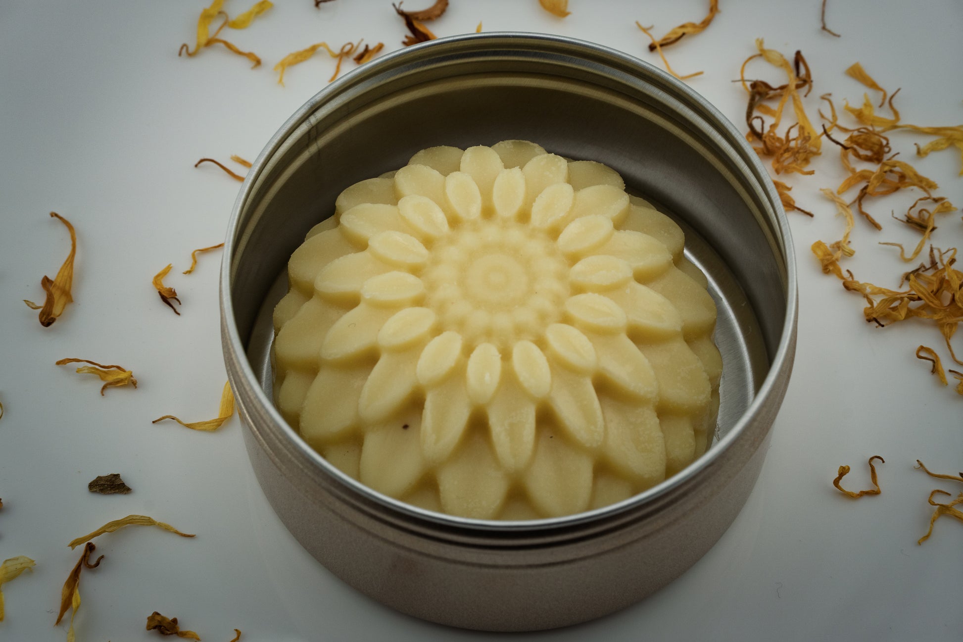 Flower-shaped tallow lotion bar in a metal tin with dried flowers around
